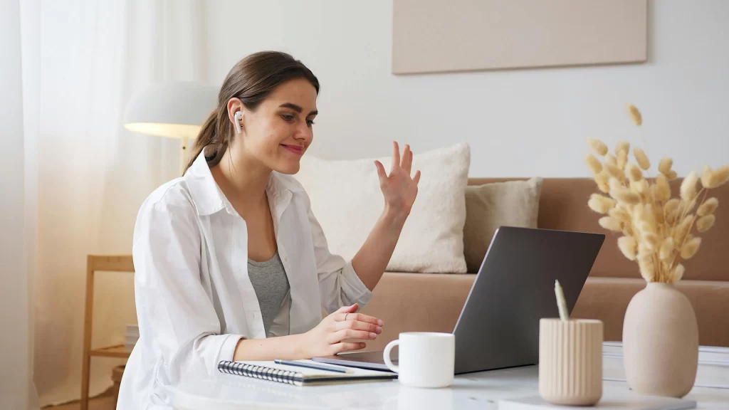 Smiling young woman wearing earphones and waving at her laptop screen during a virtual meeting, illustrating remote staffing communication.