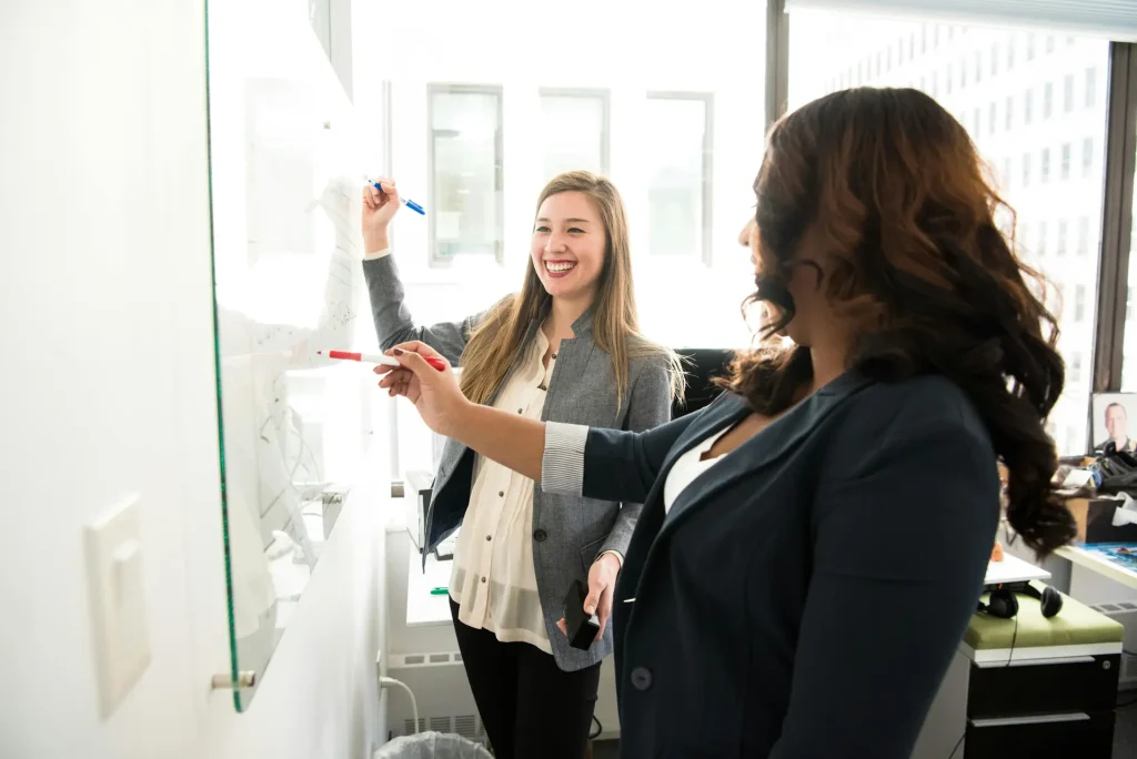 Two professional women in business attire collaborating and writing on a whiteboard during a meeting.